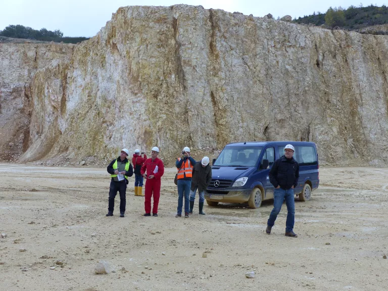 Mehrere Geologen stehen in einem Steinbruch. Im Hintergrund ist eine mehrere zehner Meter hohe Felswand.