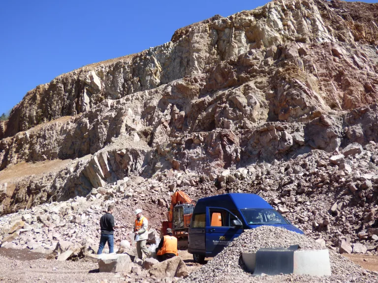 Unterhalb der Bildmitte stehen ein roter Bagger und ein blauer Transporter in einem Steinbruch. 