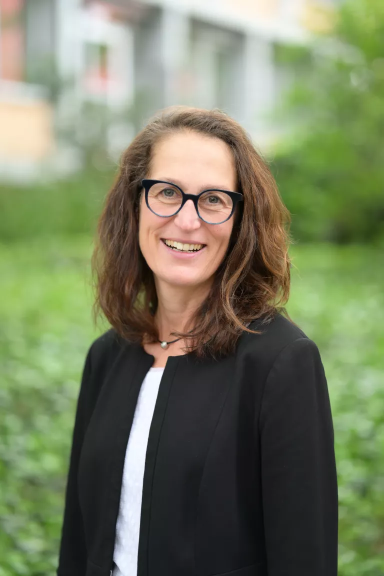 lächelnde Frau mit langen dunklen Haaren und dunkler Brille vor unscharfem Hintergrund (grüne Natur und Gebäude) 