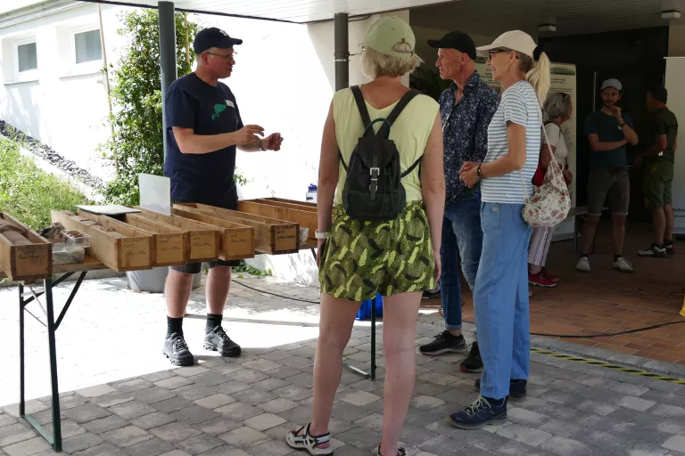 Auf einem Tisch liegen mehrere Holzkisten mit Bohrkernen; dahinter erklärt ein Mann drei zuhörenden Personen etwas
