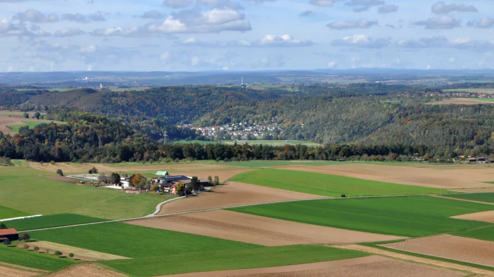 Von erhöhtem Standpunkt aus geht der Blick auf schachbrettartig angelegte Acker- und Grünlandflächen, ehe sich weiter hinten bewaldete Hänge mit bwirtschafteten Ebenen abwechseln. Noch weiter entfernt sind verschiedene Höhenzüge erkennbar.