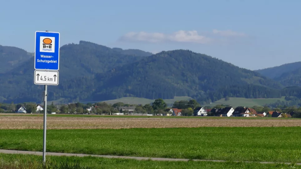 Schild mit Aufschrift "Wasserschutzgebiet". Im Hintergrund Wiesen- und Ackerflächen sowie bewaldete Hänge des Schwarzwalds.