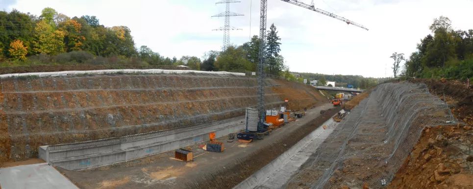 Blick auf eine Baustelle an einem länglichen Einschnitt in einem Hanggelände. In der Bildmitte sind eine im Bau befindliche Straße sowie ein Kran und weitere Baustelleneinrichtungen zu erkennen. Zu beiden Seiten der Straße ist die Hang künstlich abgetreppt. An den Stufen sind ockerfarbener bis brauner Boden und Steine zu sehen. Diese sind mit grobmaschigen Netzen abgedeckt.