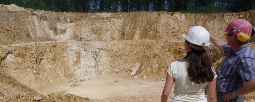 Zwei Personen stehen rechts der Bildmitte in einem Steinbruch. Die rechte Person zeigt auf den gegenüberliegenden Abbau.